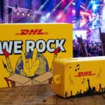 Mini câmera amarela DHL “WE ROCK” sobre mesa em palco do Rock in Rio, com público ao fundo.