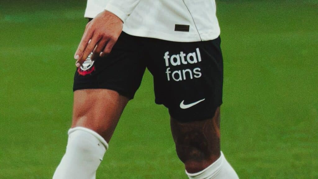 Nike e “fatal fans” nos shorts do Corinthians Jogador em campo com shorts pretos da Nike e logo “fatal fans”, em contexto de parceria com o Corinthians.