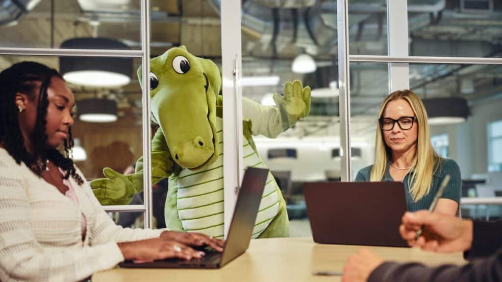 Mascote Niles da Crocs em escritório Mascote Niles, dinossauro verde, ao lado de pessoas em escritório com notebook e documentos.
