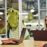 Mascote Niles, dinossauro verde, ao lado de pessoas em escritório com notebook e documentos.
