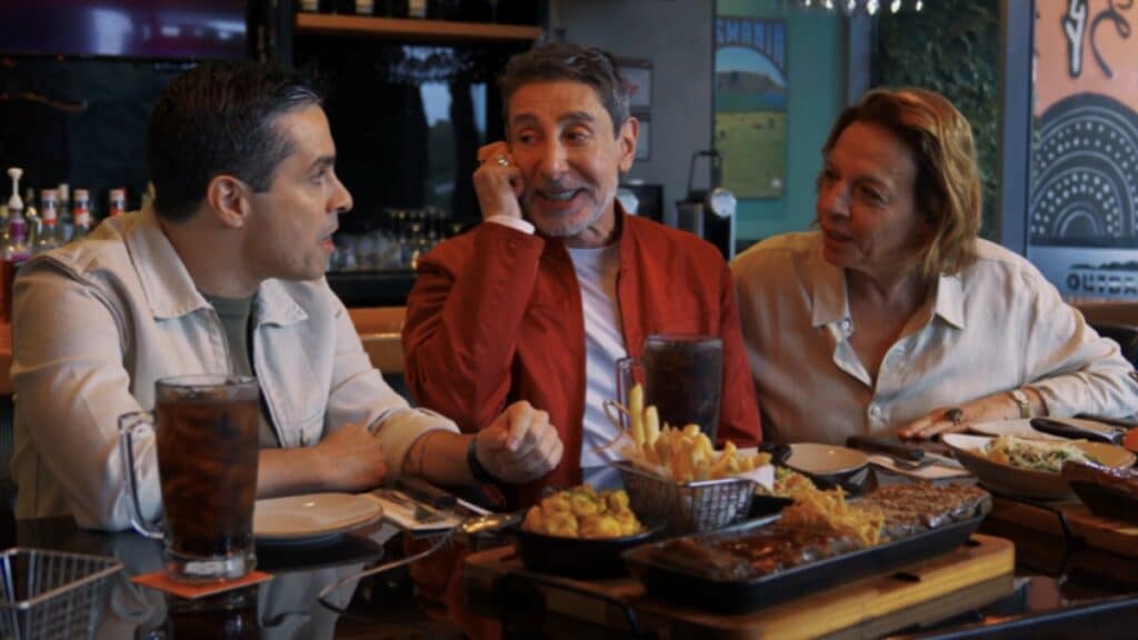 Outback Boards: amigos se reunindo para marcar o rolê Três amigos sentados em um bar/restaurante, brindando e conversando à mesa, com pratos e bebidas ao fundo.