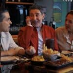 Três amigos sentados em um bar/restaurante, brindando e conversando à mesa, com pratos e bebidas ao fundo.