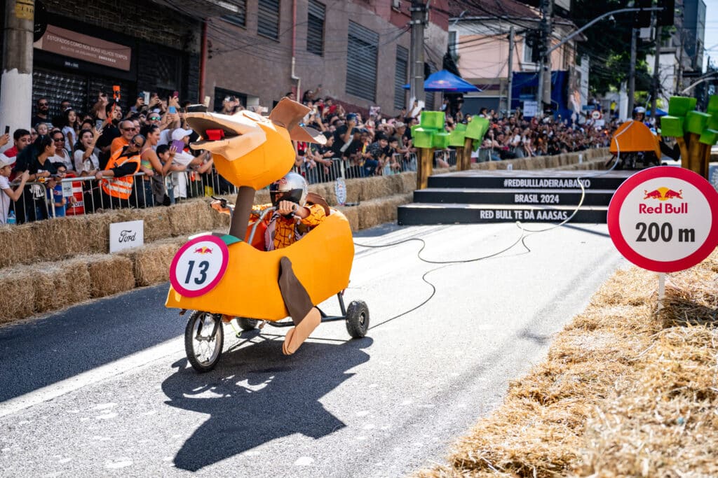 Participantes em uma corrida de carrinhos descendo uma ladeira, com placas Red Bull e público nas laterais