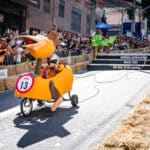 Participantes em uma corrida de carrinhos descendo uma ladeira, com placas Red Bull e público nas laterais