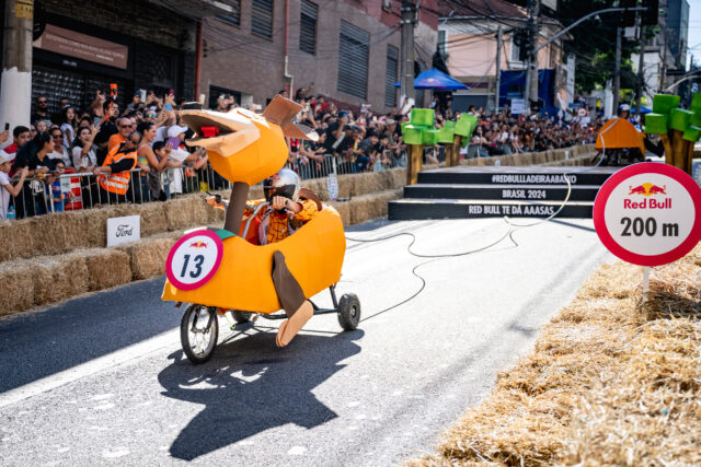 Participantes em uma corrida de carrinhos descendo uma ladeira, com placas Red Bull e público nas laterais