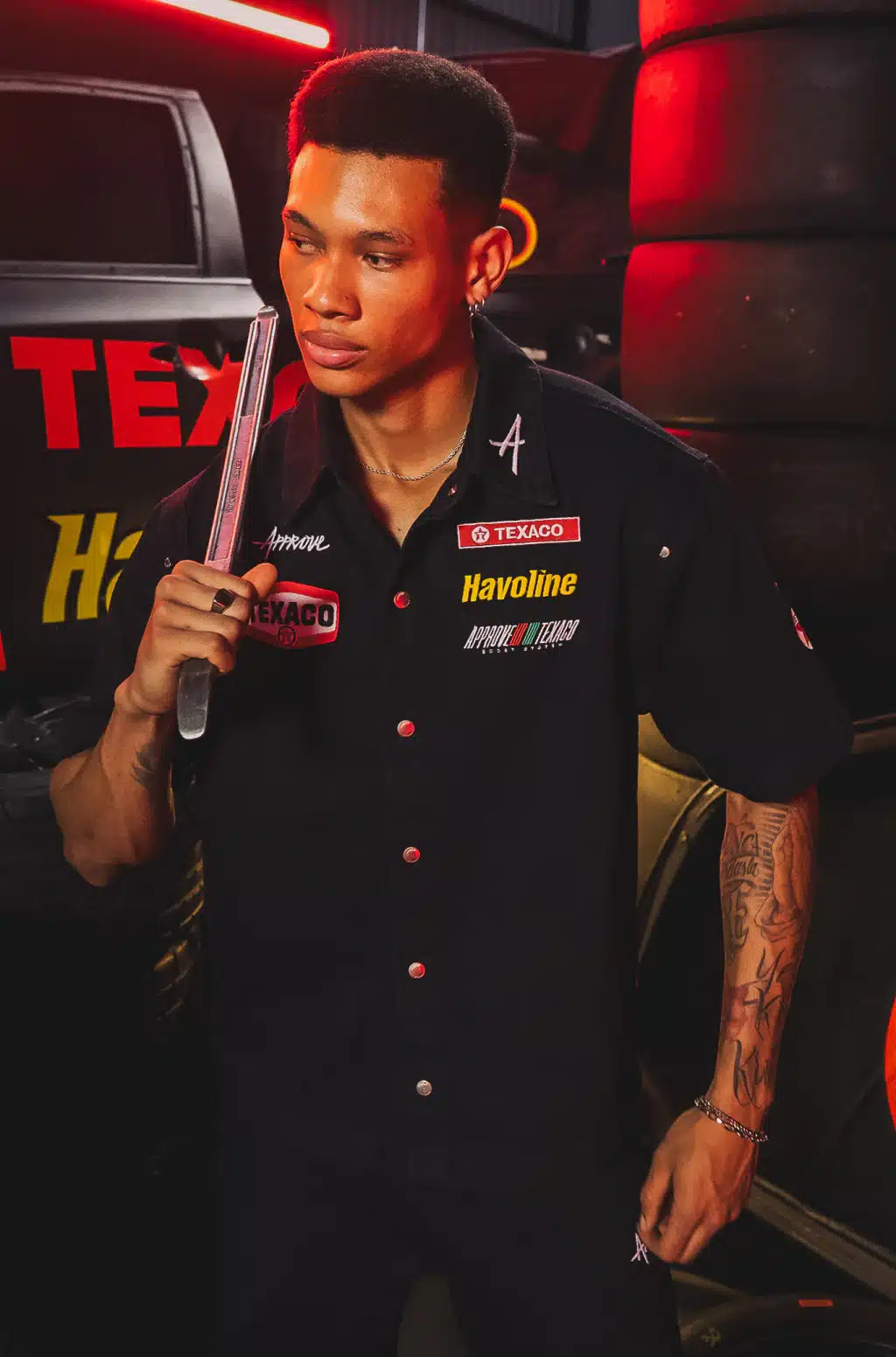 Modelo com camisa preta da Texaco x Approve em cenário com placas vermelhas, segurando uma peça metálica