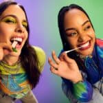 Duas mulheres sorrindo e fazendo pose com um objeto verde e branco na boca, em clima de evento ao fundo roxo e azul.