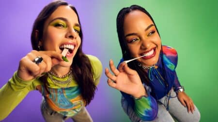 Duas mulheres sorrindo e fazendo pose com um objeto verde e branco na boca, em clima de evento ao fundo roxo e azul.