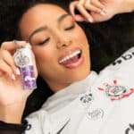 Mulher sorrindo com batom líquido da Vizzela e camisa do Corinthians, em campanha de parceria com as Brabas