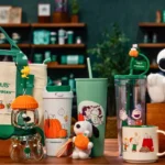 Coleção Starbucks Snoopy com Peanuts: itens e acessórios na mesa Coleção Starbucks Snoopy com itens exibidos em mesa, incluindo copos e sacola verde com personagens Peanuts.