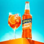 Garrafa de Aperol Spritz pronto para beber e taça com bebida alaranjada sobre fundo azul