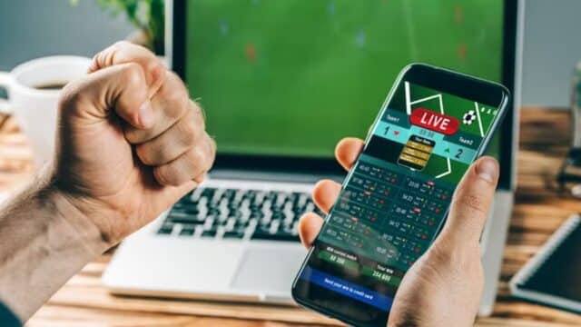Pessoa segura um celular com aplicativo de apostas enquanto aposta em mesa, em cenário esportivo e de bets