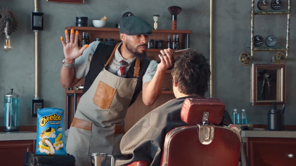 Campanha Cheetos e pó laranja nos dedos Homem usando luvas e máscara em cozinha, ao lado de embalagem de Cheetos, levantando a mão com pó laranja