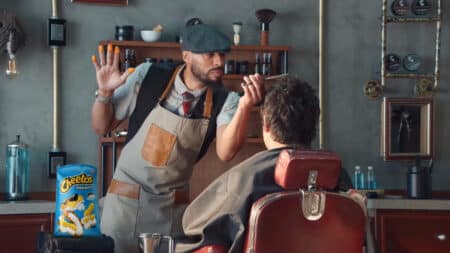 Campanha Cheetos e pó laranja nos dedos Homem usando luvas e máscara em cozinha, ao lado de embalagem de Cheetos, levantando a mão com pó laranja