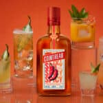 Garrafa do licor Cointreau Spicy com coquetéis ao lado, em fundo laranja, com pimenta e pepino como decoração