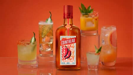 Cointreau Spicy e coquetéis com pimenta e pepino Garrafa do licor Cointreau Spicy com coquetéis ao lado, em fundo laranja, com pimenta e pepino como decoração