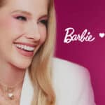Modelo sorrindo usando joias e brincos, em campanha da collab Barbie x Misha, com fundo rosa.