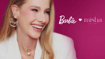 Barbie x Misha: joias inspiradas na Barbie em campanha Modelo sorrindo usando joias e brincos, em campanha da collab Barbie x Misha, com fundo rosa.