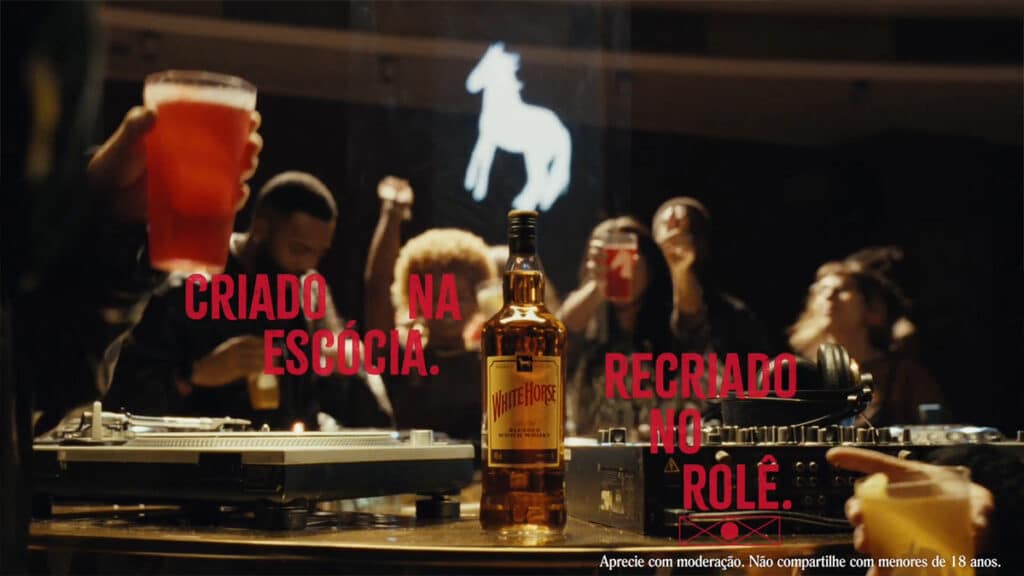 Garrafa de uísque White Horse em cena noturna com pessoas ao fundo e letreiro “Criado na Escócia” e “Recriado no rolê”.