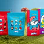 Embalagens de chocolate Kopenhagen com tema Smurfs, com personagens dos Smurfs, sobre grama e cogumelos coloridos ao fundo.