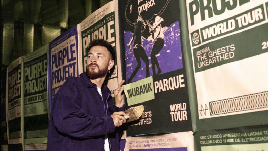 Fred em evento com banners Nubank Parque Homem em palco segurando papel ao lado de banners com a marca Nubank Parque, durante evento do Purple Project World Tour.