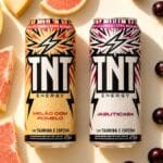Latas de TNT Energy Drink sabores Melão com Pomelo e Jabuticaba com porções de frutas ao fundo.