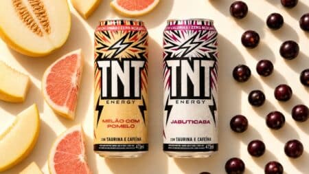 TNT Energy Drink Jabuticaba e Melão com Pomelo Latas de TNT Energy Drink sabores Melão com Pomelo e Jabuticaba com porções de frutas ao fundo.