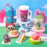Mesa com bebidas e sobremesas temáticas das Meninas Superpoderosas da We Coffee, com copos coloridos e enfeites em fundo azul e verde.