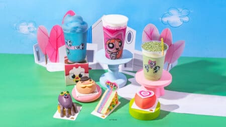 Mesa com bebidas e sobremesas temáticas das Meninas Superpoderosas da We Coffee, com copos coloridos e enfeites em fundo azul e verde.