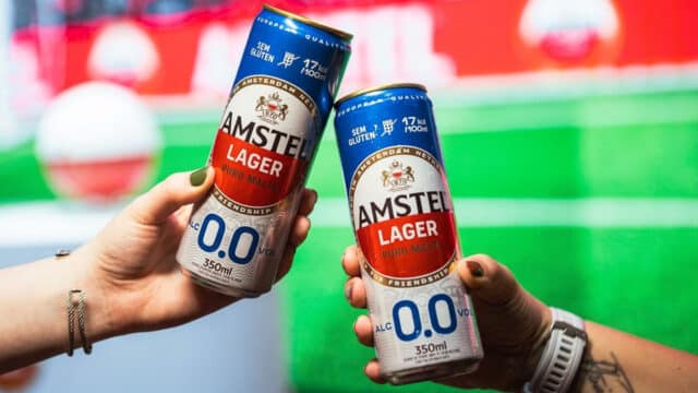 Duas latas de Amstel Lager 0.0 zero álcool e zero glúten em mãos, com fundo colorido verde e vermelho