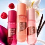 Frascos de body splash e hair & body mist La Vie Est Belle da Lancôme com flores em fundo claro.