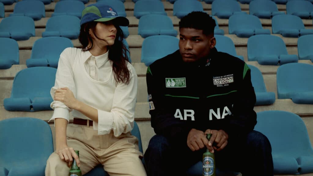 Heineken 0.0 e Carnan: foto na arquibancada Duas pessoas sentadas em arquibancada azul, uma ao lado de uma Heineken 0.0, em foto promocional da coleção Carnan.
