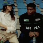 Duas pessoas sentadas em arquibancada azul, uma ao lado de uma Heineken 0.0, em foto promocional da coleção Carnan.