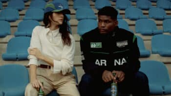 Heineken 0.0 e Carnan: foto na arquibancada Duas pessoas sentadas em arquibancada azul, uma ao lado de uma Heineken 0.0, em foto promocional da coleção Carnan.