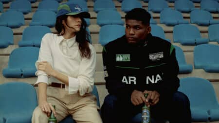 Heineken 0.0 e Carnan: foto na arquibancada Duas pessoas sentadas em arquibancada azul, uma ao lado de uma Heineken 0.0, em foto promocional da coleção Carnan.