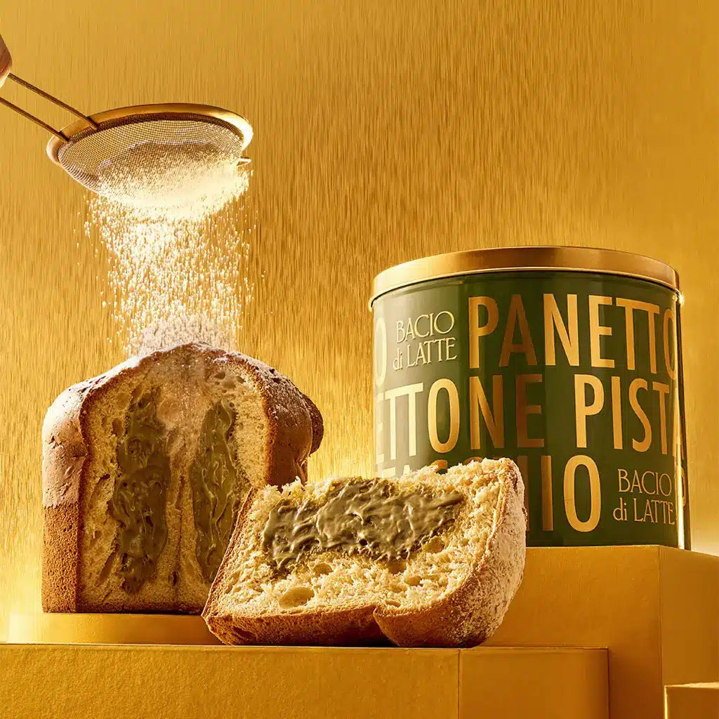 Panettone Bacio di Latte com fatias abertas e lata TONE PISTACIO sobre bancada com fundo dourado