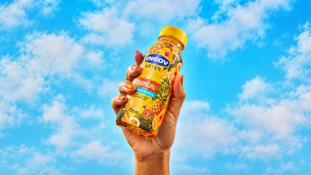 Engov After Tropical: edição limitada para o verão Mão segurando embalagem de Engov After sabor Tropical, com fundo de céu azul e nuvens.