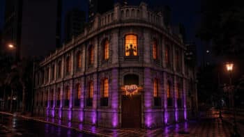 Fábrica Assombrada da Fanta com fachada iluminada Fachada iluminada da Fábrica Assombrada da Fanta à noite, com luzes roxas e janela em clima de Halloween