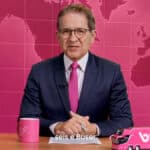 Carlos Tramontina em telejornal humorístico Seis e Buser, com fundo em mapa e objetos de ônibus sobre a mesa