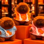 Três capivaras de pelúcia com moletom em tons azul, laranja e rosa, em expositores com ambiente de loja ao fundo.