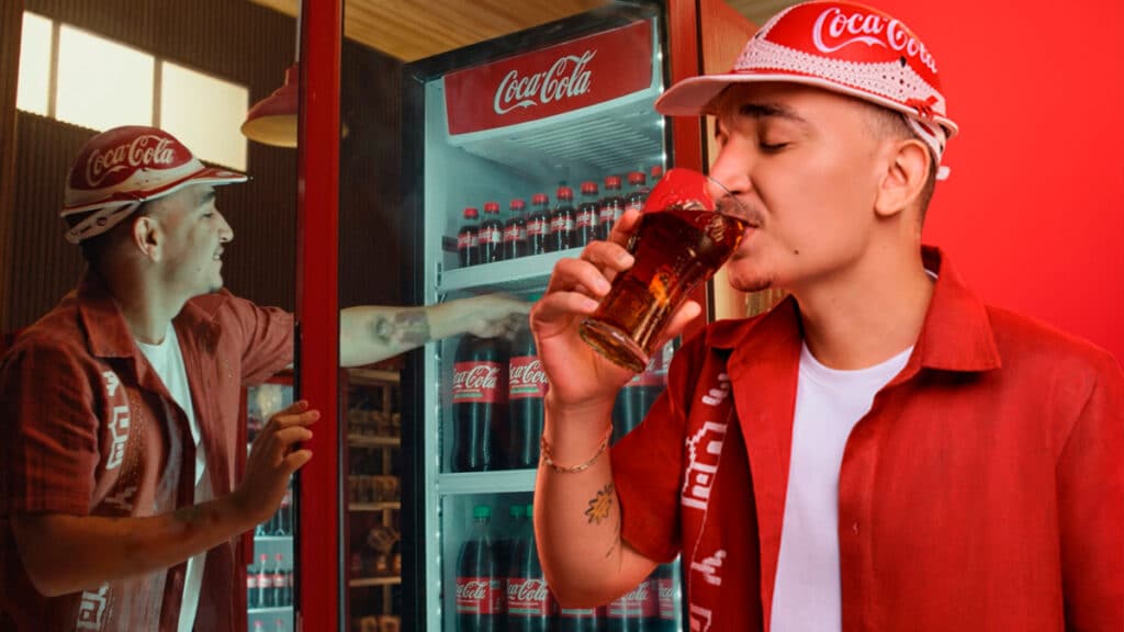João Gomes em campanha da Coca-Cola, brindando com garrafas retornáveis em um estabelecimento.