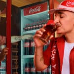 João Gomes em campanha da Coca-Cola, brindando com garrafas retornáveis em um estabelecimento.