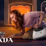 Campanha de Natal da Prada com mulher deitada ao lado de bolsa e cenário iluminado com luz laranja ao fundo.