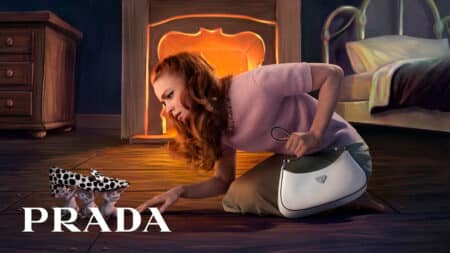 Campanha de Natal 2026 da Prada com ilustração e bolsa Campanha de Natal da Prada com mulher deitada ao lado de bolsa e cenário iluminado com luz laranja ao fundo.