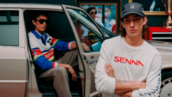 Modelo posa ao lado de carro usando boné e camiseta branca com o logo “SENN A”, em campanha inspirada em Ayrton Senna.