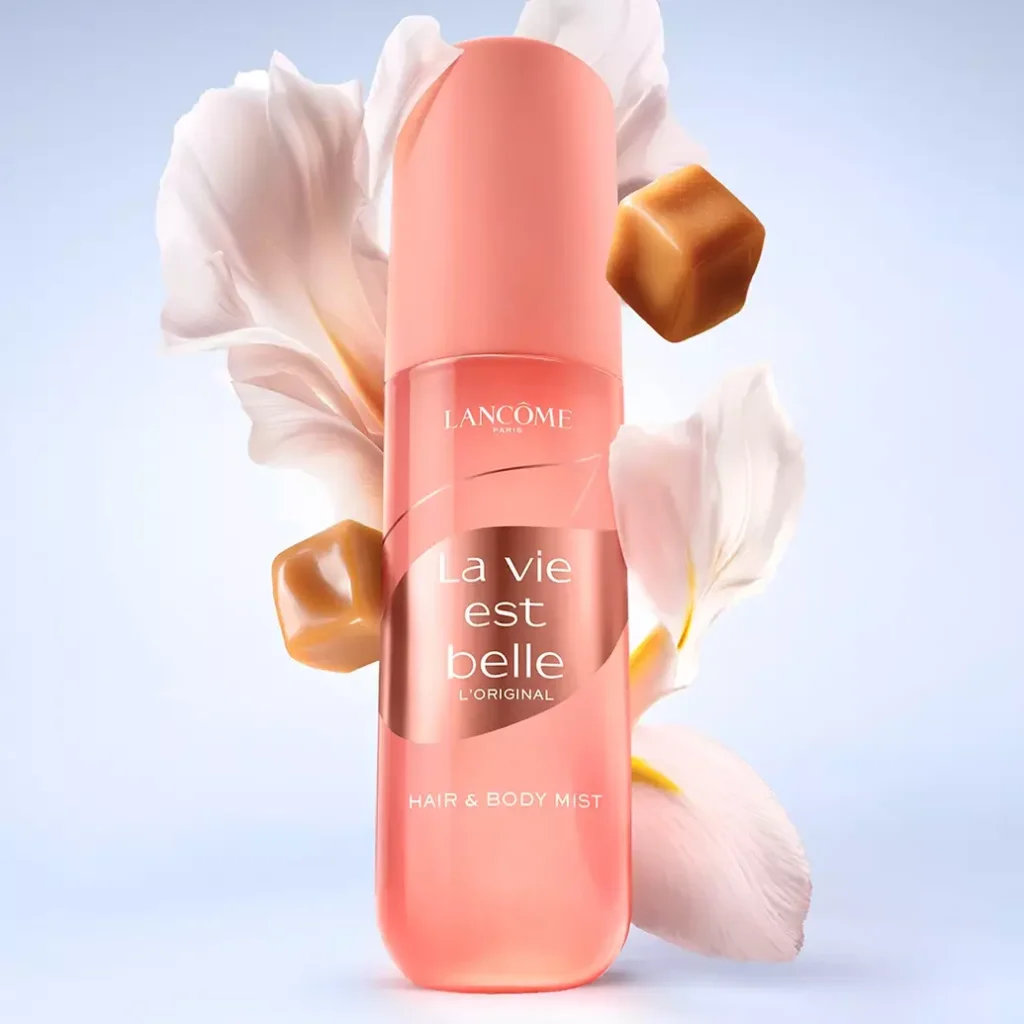 Frasco de body splash La Vie Est Belle L’Original da Lancôme, com embalagem rosa e fundo claro, ao lado de pétalas