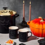 Panelas e xícaras temáticas de Halloween da Le Creuset sobre mesa, com abóbora laranja e velas acesas