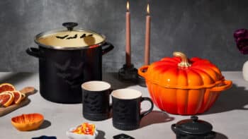 Le Creuset Halloween: panelas e abóbora temática na mesa Panelas e xícaras temáticas de Halloween da Le Creuset sobre mesa, com abóbora laranja e velas acesas