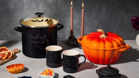 Le Creuset Halloween: panelas e abóbora temática na mesa Panelas e xícaras temáticas de Halloween da Le Creuset sobre mesa, com abóbora laranja e velas acesas