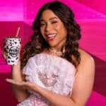Mulher sorrindo em cenário rosa segurando um copo da Milky Moo com estampa de vaquinhas, usando vestido claro e colar de pérolas.